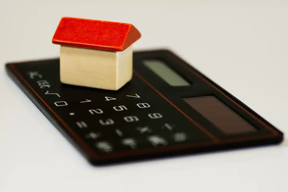 Empty mortgage application forms and a calculator on a desk, with a rising stock chart displayed on a nearby screen, suggesting financial strain and market volatility.