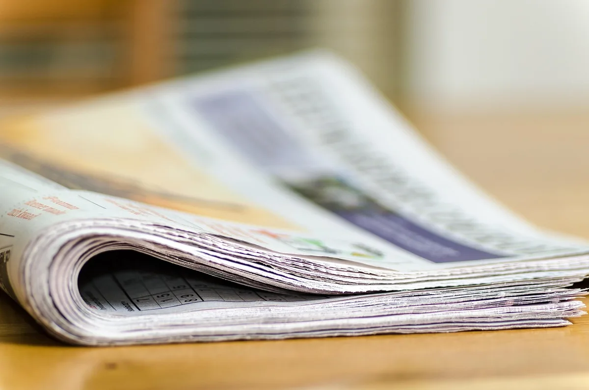 Close-up of hands leafing through aged newspaper pages in an archive box, typed headlines visible but unreadable