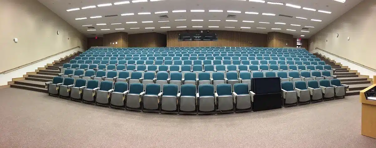 A lone anonymous student in a classroom looking at an empty enrollment screen, hands on a desk, a closed textbook beside them — tension