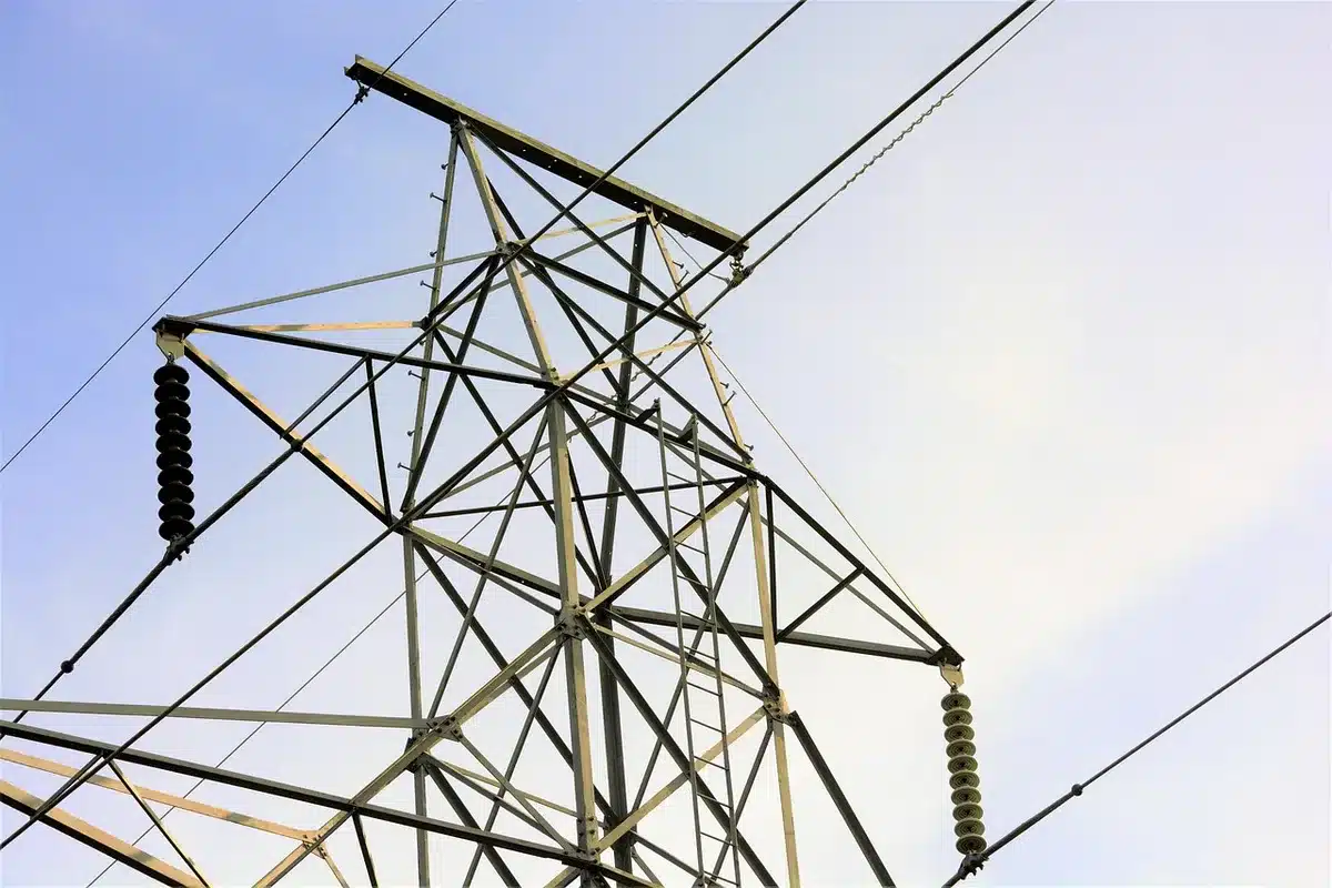 An empty transmission tower against a cloudy sky, with power lines stretching into the distance, suggesting grid infrastructure and vulnerability.