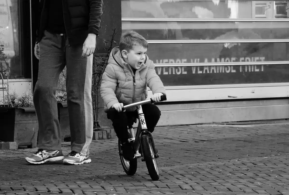 A soft-focus scene of an empty family garden path with a small unbranded child’s bicycle leaning on a fence and a pair of tiny shoes on the grass