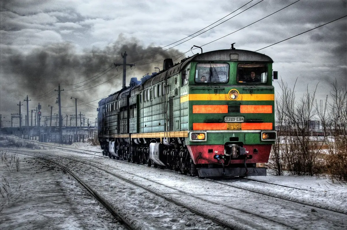 A long passenger train stopped on tracks with doors open and anonymous staff guiding evacuated passengers away, a few bags on the platform and emergency vehicles in the distance