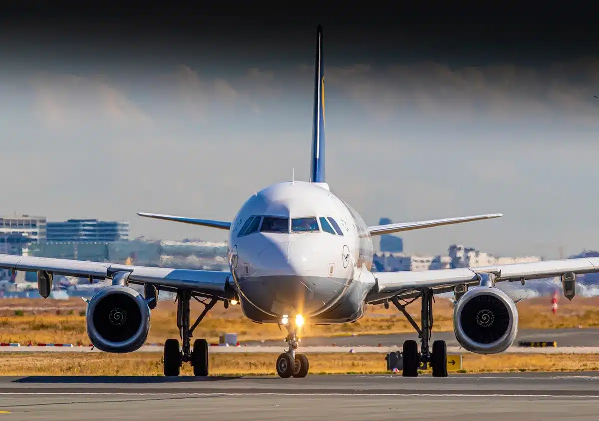 A commercial aircraft on a tarmac with departure gates visible in the background, a clear sky above, and ground crew equipment idle