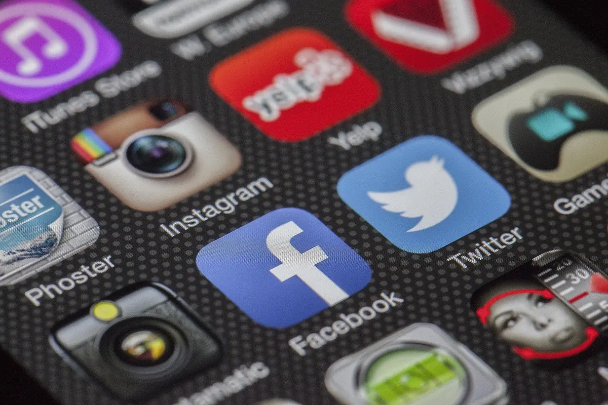 A dimly lit anonymous smartphone held over a dark table showing a blurred social feed with unreadable text, a discarded campaign button beside it, suggesting controversy over online posts