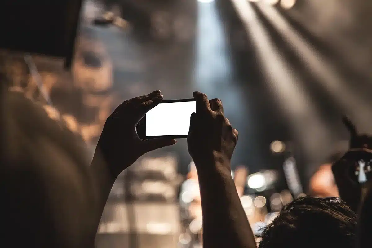 A packed theater crowd with silhouettes of raised hands and heads in shadow, cinema seats stretching into darkness, warm light from a distant screen illuminating anonymous faces