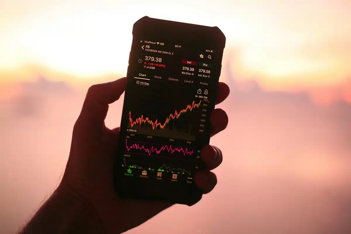 A computer monitor displaying a stock market index chart with upward trend lines in muted blues and greens, a child's piggy bank silhouette in soft focus beside it, representing long-term childhood investment growth.
