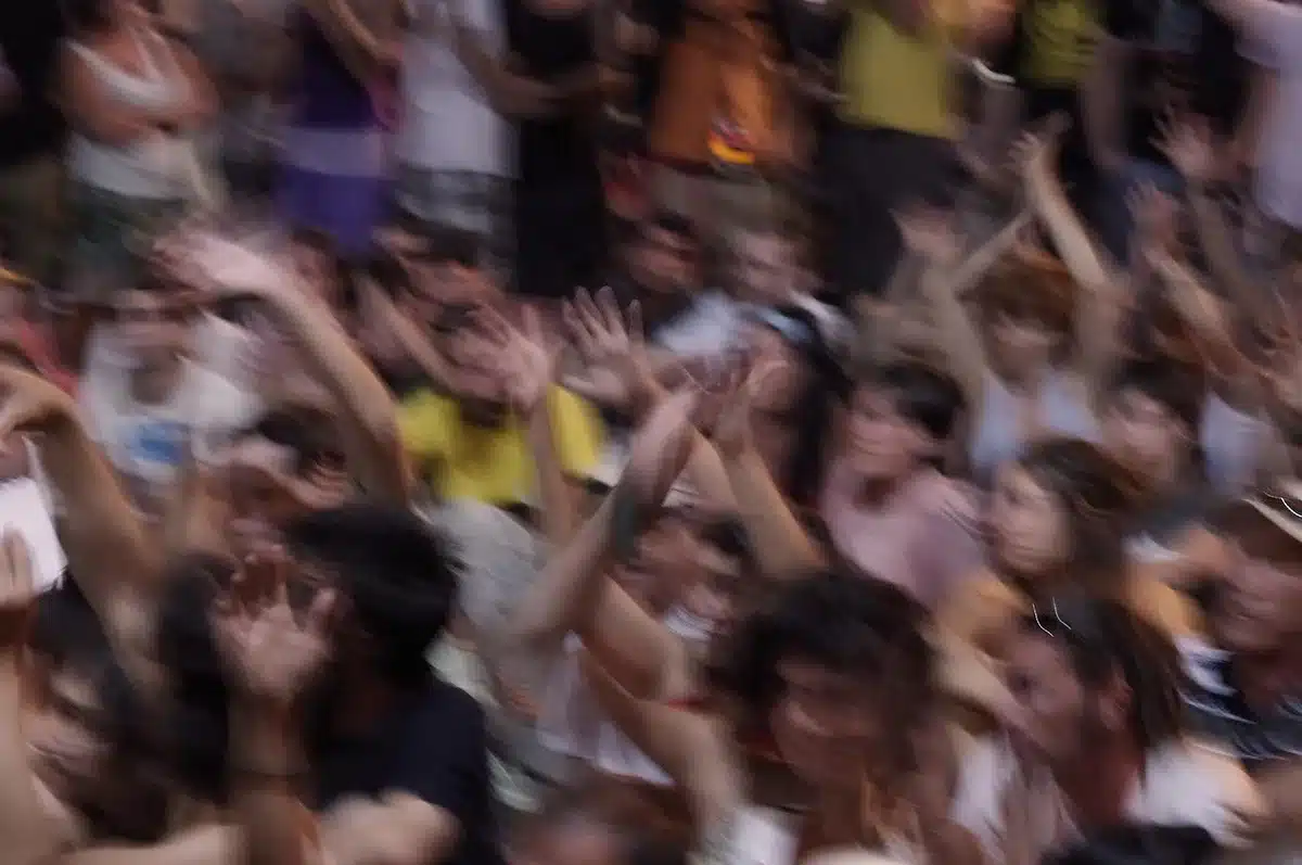 Anonymous crowd of supporters seen from behind, a sea of raised hands and blurred signs under arena lights, faces indistinct in motion blur