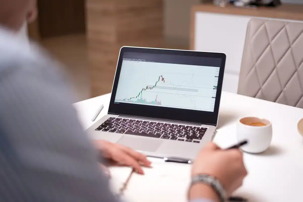 close-up of a laptop screen showing a generic stock chart with TSLA ticker blurred, an anonymous hand beside a notebook