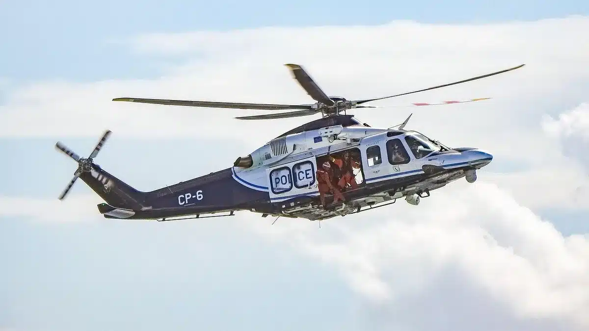 Distant emergency crews and a burned rotorcraft wreck in a small commercial-carpark, smoke rising with nearby damaged vehicles and storage containers