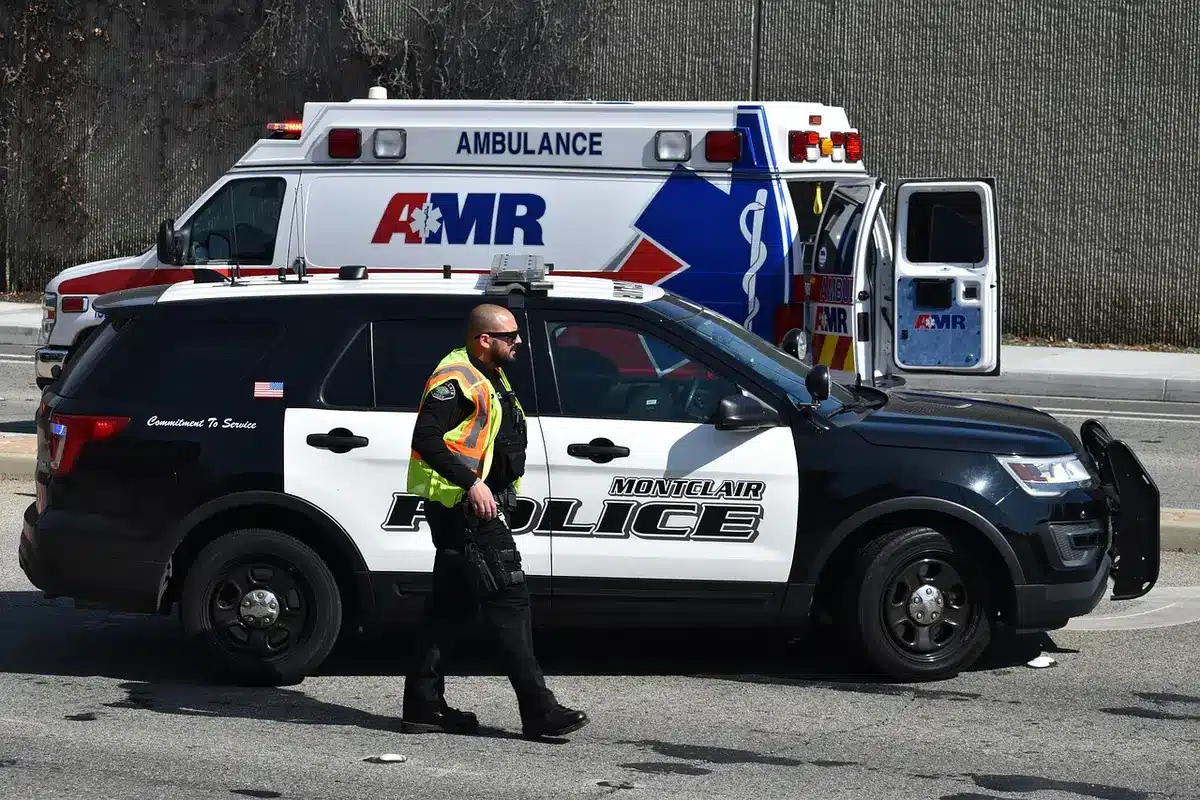 Anonymous first responders and a stretcher being moved past a low commercial building and burned cars, scene cordoned by emergency tape