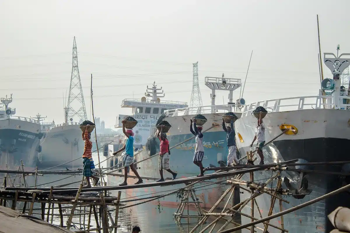 Port container stacks and cargo ships at a border crossing facility, customs checkpoint with barriers and warning signs, overcast sky emphasizing economic uncertainty
