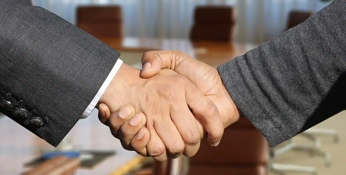 hands on a contract document on a wood table, pen beside unsigned pages, a laptop screen in soft focus