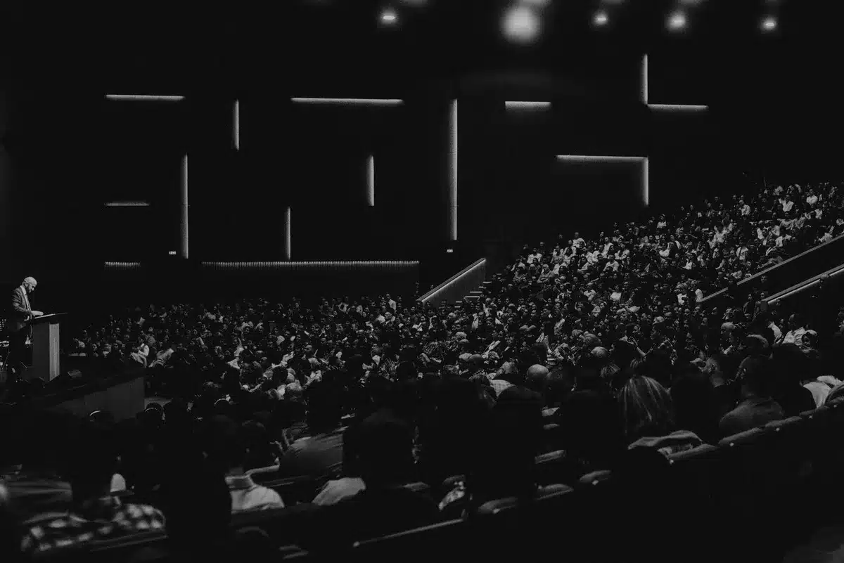 Wide symbolic scene of an empty lectern, a single microphone on a stand under a spotlight on a stage, dust motes in the beam and an American-flag suggestion out of focus — anticipation