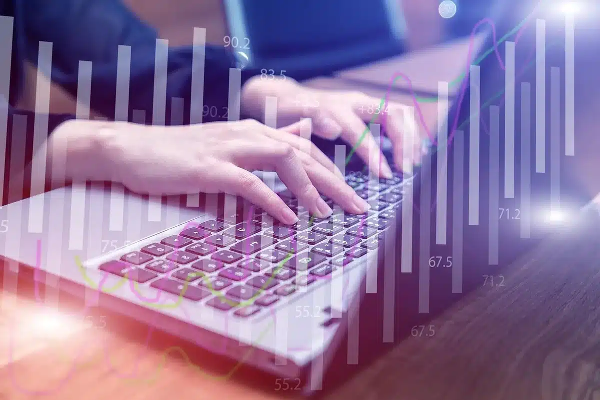 anonymous hands on a keyboard next to a generic FX chart on a laptop screen, blurred city window behind