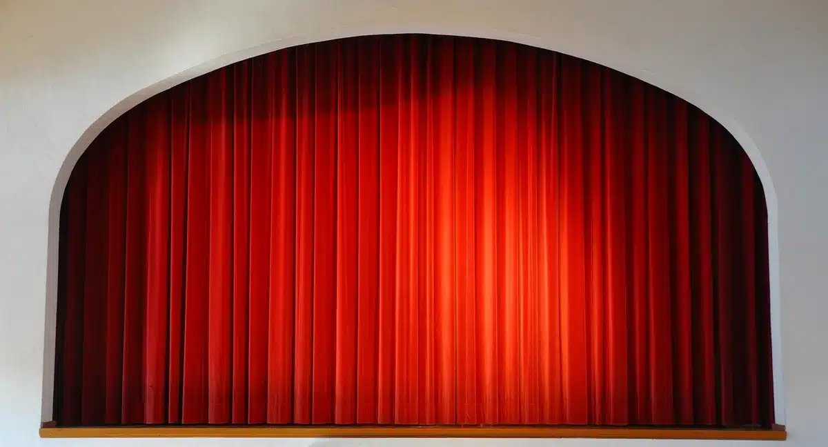 An empty campaign stage with a single microphone on a stand under one spotlight, the backdrop blurred and seats in the dim auditorium visible — anticipation before a political event