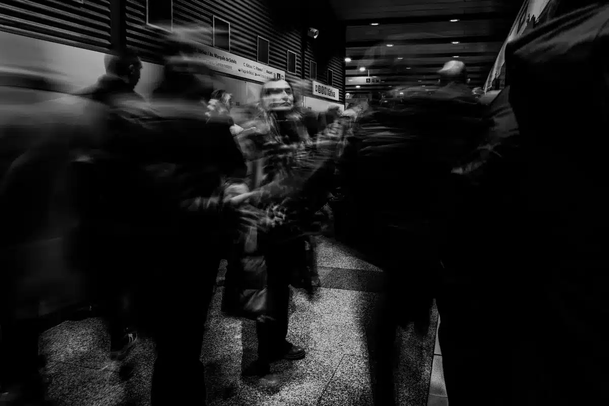 A small crowd of anonymous supporters gathered outside a generic civic hall, flags and signs blurred, people shown only as silhouettes in late-afternoon light — pre-event gathering mood
