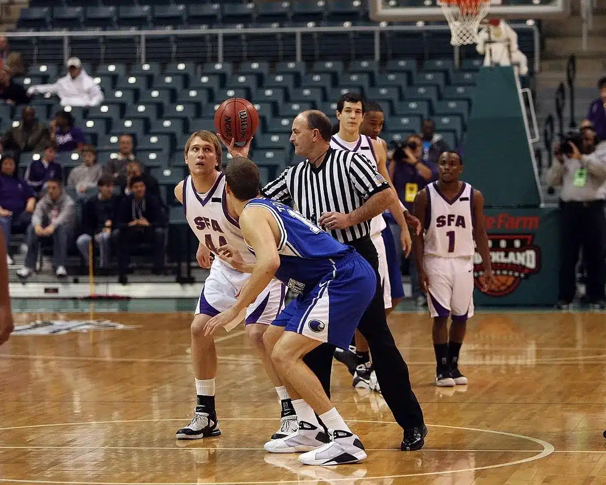 A close-up of a folded college basketball jersey on a wooden bench, no logos visible, an old scoreboard blurred in the background