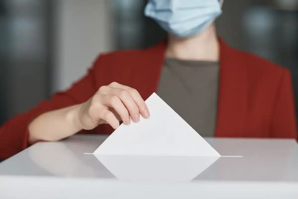A single empty polling station table with an unbranded ballot box and stapled information sheets, a lone pen resting nearby suggesting imminent voting activity