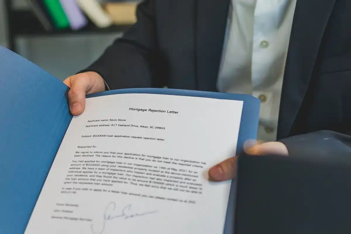 An anonymous desk with a stack of unbranded mortgage documents, a generic pen resting on top, an out-of-focus laptop screen showing a generic line chart in the background
