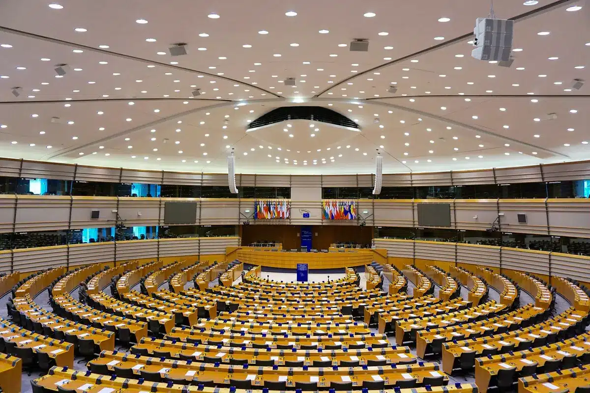 An empty assembly chamber with rows of vacant desks and a single microphone on a desk centre-frame, papers scattered, a folded resolution page visible but unreadable