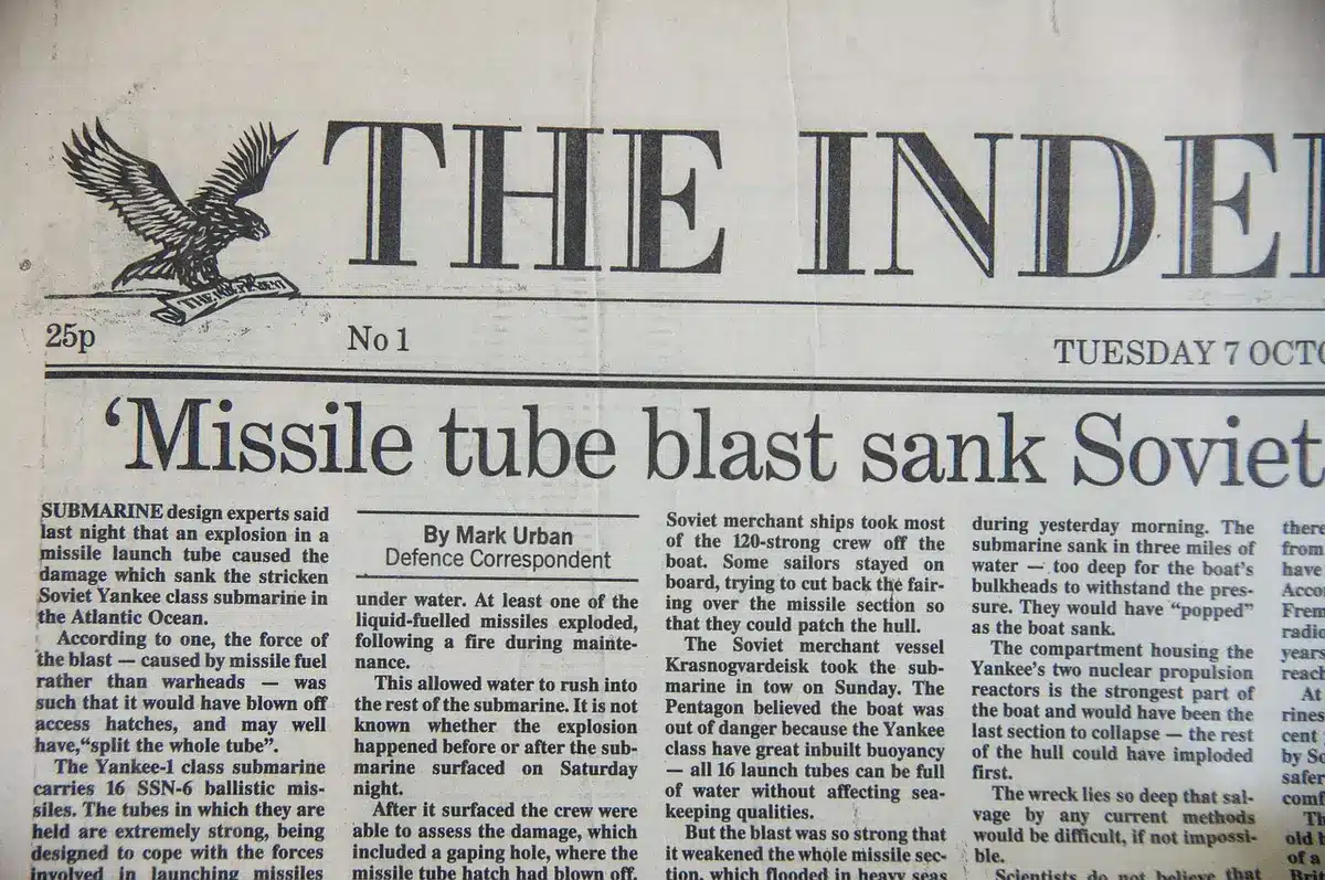 a close-up of a printed page headlined by a 1980s date, folded on a wooden table with an anonymous pair of reading glasses nearby