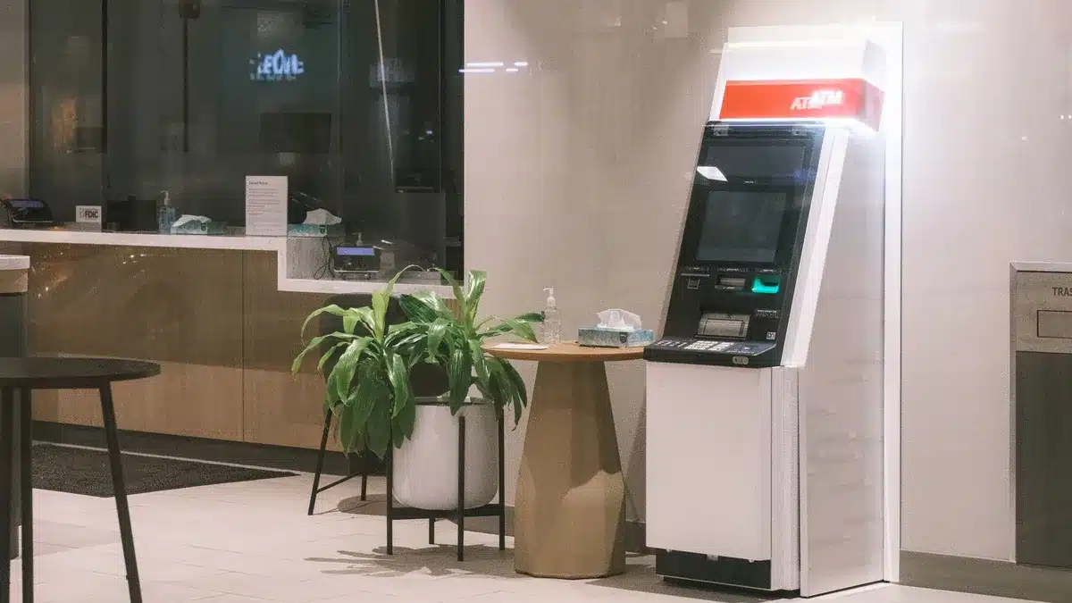 A generic bank counter with an anonymous customer speaking to a teller, a posted calendar visible behind the counter showing late October dates, no logos or identifiable staff