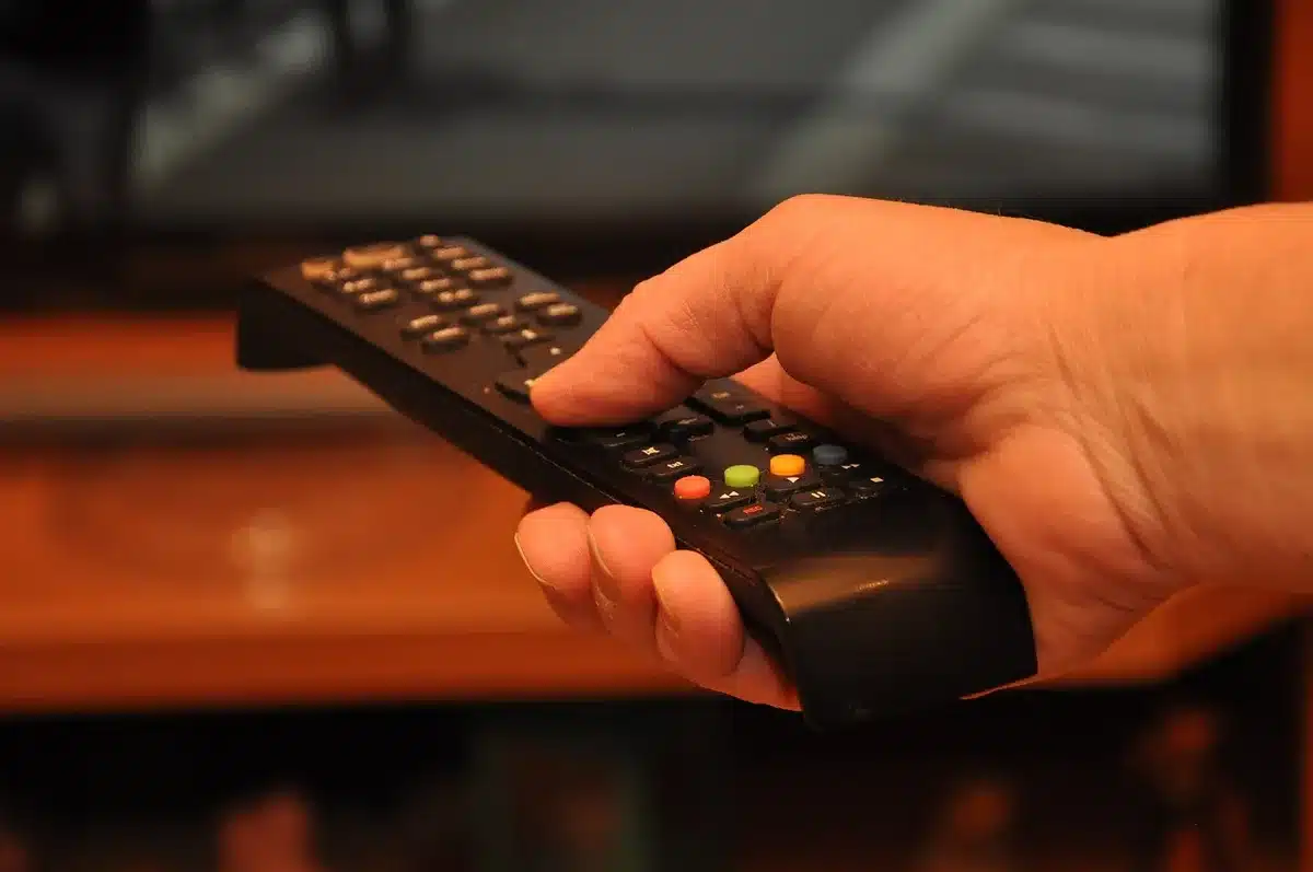 An anonymous hand holding a turned-off television remote in front of a dark living room, suggesting viewers deciding whether to tune into a controversial ad